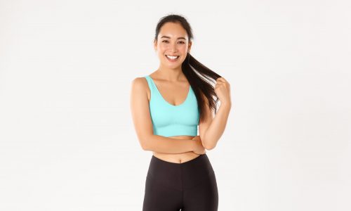 Sport, wellbeing and active lifestyle concept. Smiling pretty asian girl in sportswear, playing with ponytail and looking happy at camera after good gym workout, standing white background.
